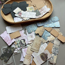 A collection of vintage-style paper craft embellishments, including die-cut labels, stamps, and frames, arranged on a gray surface with a wooden tray in the background.