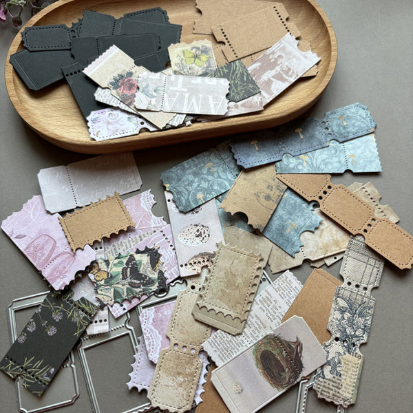 A collection of vintage-style paper craft embellishments, including die-cut labels, stamps, and frames, arranged on a gray surface with a wooden tray in the background.