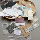 A flat lay of scrapbooking paper sheets, embossed cardstock pieces, and metal die-cut frames arranged on a gray surface with a wooden tray in the background.