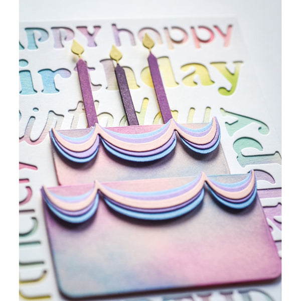 A close-up of a pastel birthday card featuring a layered cake design with purple candles, surrounded by embossed lettering on a white cardstock background.