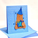 A handmade 3D pop-up birthday card featuring a brown felt teddy bear wearing a blue polka-dot party hat and holding a blue and green gift box, set against a light blue embossed card background with 'Happy Birthday' text.