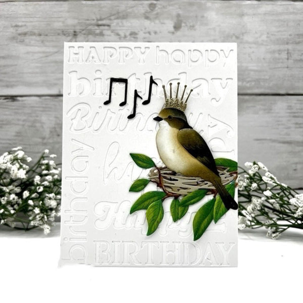 A handmade white birthday card featuring a bird perched on a nest with green leaves, a small gold crown, and black musical notes, set against a rustic gray wood background with white flowers on either side.