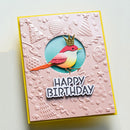 A handmade square birthday card with a pink embossed background, a yellow border, and a central circular cutout featuring a colorful bird wearing a gold crown above the words “HAPPY BIRTHDAY.”.