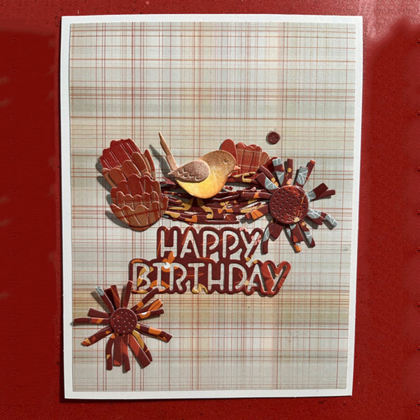 A handmade layered paper card with a plaid background, featuring a small bird, autumn leaves, and 3D text that reads 'HAPPY BIRTHDAY' in brown letters, set against a solid red background.