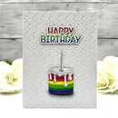 A square white greeting card with a textured dotted pattern, featuring the words 'HAPPY BIRTHDAY' in rainbow colors above a small rainbow-striped birthday cake with dripping white frosting. The card is displayed on a light gray wood-plank surface with white roses in the background.