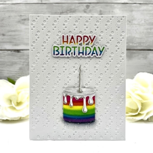 A square white greeting card with a textured dotted pattern, featuring the words 'HAPPY BIRTHDAY' in rainbow colors above a small rainbow-striped birthday cake with dripping white frosting. The card is displayed on a light gray wood-plank surface with white roses in the background.