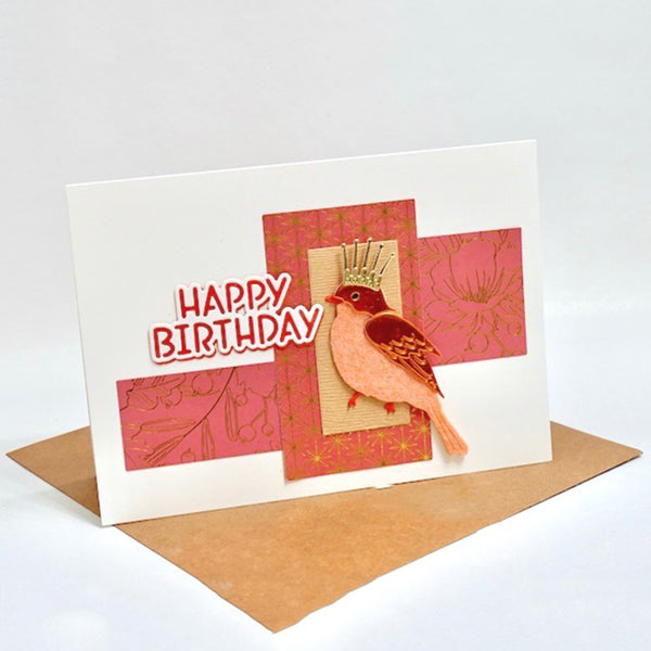 A handmade birthday card with a white base, layered pink patterned paper, a die-cut bird with a small gold crown, and the words “HAPPY BIRTHDAY” in red and white. A brown envelope is placed behind the card on a light gray background.