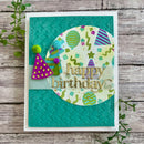 A handmade square birthday card with a white border, featuring a textured teal background, a circular patterned insert with colorful party motifs, and raised gold 'happy birthday' lettering. The card is photographed on a rustic wood surface with small green leaves around it.