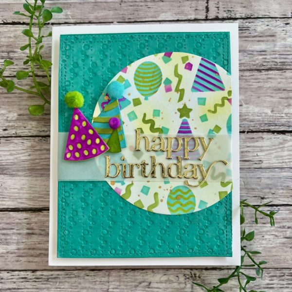 A handmade square birthday card with a white border, featuring a textured teal background, a circular patterned insert with colorful party motifs, and raised gold 'happy birthday' lettering. The card is photographed on a rustic wood surface with small green leaves around it.