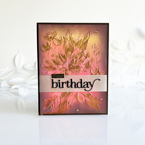 A handmade square greeting card with a floral design in pink, gold, and brown tones, featuring a white banner across the middle that reads 'Happy birthday' in black script. The card is photographed standing upright on a white surface with a soft, blurred white floral background.