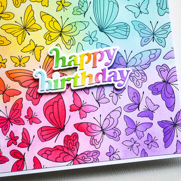 A close-up of a colorful handmade card featuring a rainbow gradient background with illustrated butterflies in red, pink, purple, blue, and yellow. A die-cut sticker reading “happy birthday” is layered over the design.