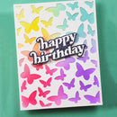 A square white greeting card with layered butterfly cutouts in yellow, green, blue, pink, orange, and purple, plus a glittery 'happy birthday' message on the front, photographed against a teal background.