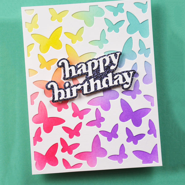 A square white greeting card with layered butterfly cutouts in yellow, green, blue, pink, orange, and purple, plus a glittery 'happy birthday' message on the front, photographed against a teal background.
