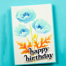 A handmade-style birthday card featuring three light blue watercolor poppy flowers with orange and yellow leaves on a white cardstock, with 'happy birthday' in black script with white outlines. The card is photographed standing upright against a bright turquoise-blue textured background.