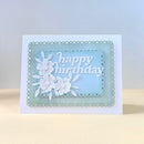 A handmade square birthday card with a white base, featuring a layered light blue and mint green scalloped frame, white floral cutouts, and the words 'happy birthday' in white script.