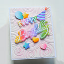 A square handmade birthday card with a white embossed swirl background and colorful layered lettering that reads 'Happy Birthday.' Decorative elements include pastel hearts, stars, and a striped party hat.