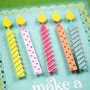 A close-up of a handmade birthday card featuring five paper candle decorations in yellow, pink, orange, and blue, with the text 'make a' visible on a teal card base.