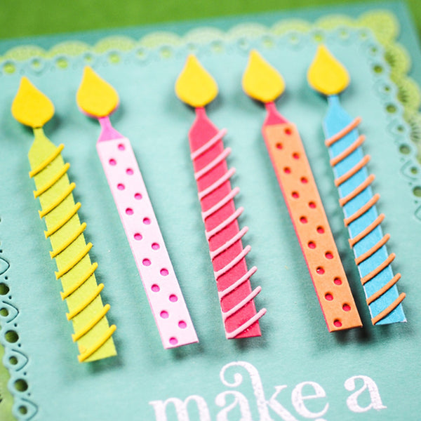 A close-up of a handmade birthday card featuring five paper candle decorations in yellow, pink, orange, and blue, with the text 'make a' visible on a teal card base.