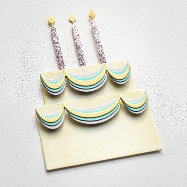 A two-tier birthday cake decoration set with pastel striped tiers and three glittery candle props on a light textured background.