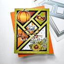 A handmade autumn-themed greeting card with a geometric diamond design, featuring a pumpkin, acorns, mushrooms, and a yellow flower, tied with a twine bow. The card is displayed on top of an orange envelope with a silver die-cutting machine visible in the background.