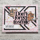 A handmade greeting card featuring two cartoon koalas, the phrase 'Don't forget to smile' in layered black and pink lettering, and a 'SAY CHEESE!' tab, displayed on a light gray wood background.