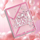 A pink handmade birthday card with 'Happy Birthday' in glittery pink lettering, decorated with a bow motif, sequins, and a clear window, shown against a background of pink balloons.