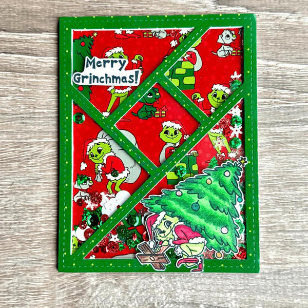 A square Christmas card featuring a green border and a red background printed with Grinch-themed illustrations, including the phrase 'Merry Grinchmas!' on a white banner. The card is photographed flat on a light gray wood surface.