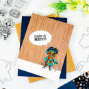 A handmade pirate-themed greeting card featuring a wood-grain pattern background, a cartoon cat in a pirate outfit, and a speech bubble that reads 'THANK YE MATEY!'. The card is layered over navy blue and kraft paper envelopes on a white flat-lay surface with craft cutters and flowers.