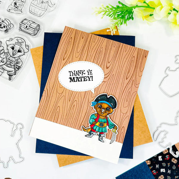 A handmade pirate-themed greeting card featuring a wood-grain pattern background, a cartoon cat in a pirate outfit, and a speech bubble that reads 'THANK YE MATEY!'. The card is layered over navy blue and kraft paper envelopes on a white flat-lay surface with craft cutters and flowers.