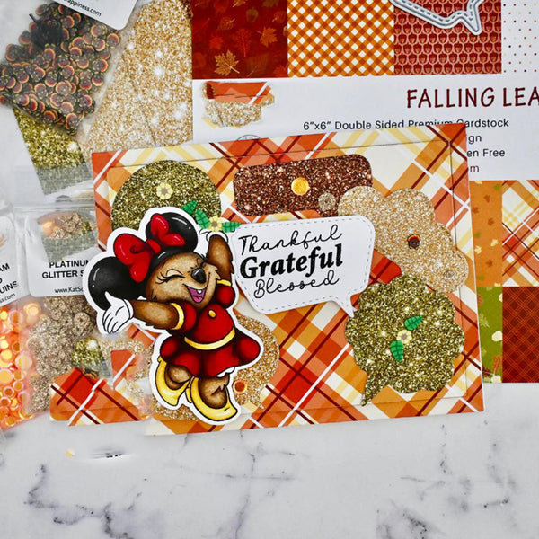 A styled flat-lay of craft cardstock sheets and glitter bags featuring autumn fall themes, including orange plaid, red and orange checkered patterns, and gold glitter. A Minnie Mouse sticker with the text 'Thankful Grateful Blessed' is placed on top of the cardstock.