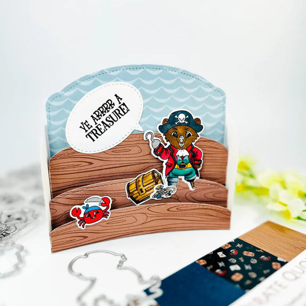 A handmade pirate-themed card featuring a cartoon bear dressed as a pirate, a treasure chest, a red crab, and a speech bubble that reads 'VE LOST A TREASURE'. The card is styled on a white surface with craft dies and a patterned card partially visible in the foreground.