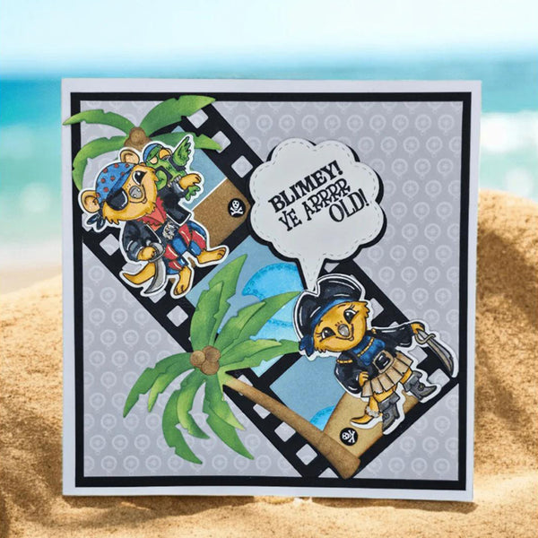 A square handmade greeting card with a pirate-themed cartoon bear design, placed on a sandy beach with a blurred ocean background. The card features a gray patterned background, a black border, a film strip motif, and a speech bubble that reads 'BLIMEY VE KARAĞAZI OLDU!'.