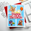 A square handmade birthday card featuring cartoon bears, a party hat, balloons, presents, and cupcakes on a light blue background with large red and white 'Happy Birthday' text. The card is styled on red envelopes with metal die-cutting and scoring tools nearby.