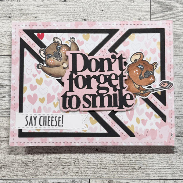 A handmade greeting card featuring two cartoon koalas, the phrase 'Don't forget to smile' in layered black and pink lettering, and a 'SAY CHEESE!' tab, displayed on a light gray wood background.