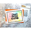 A handmade greeting card with the phrase 'Don't forget to smile' in black script over a pastel rainbow and floral pattern, displayed with metal die-cut embellishments and a stamp sheet on a gray marble surface.
