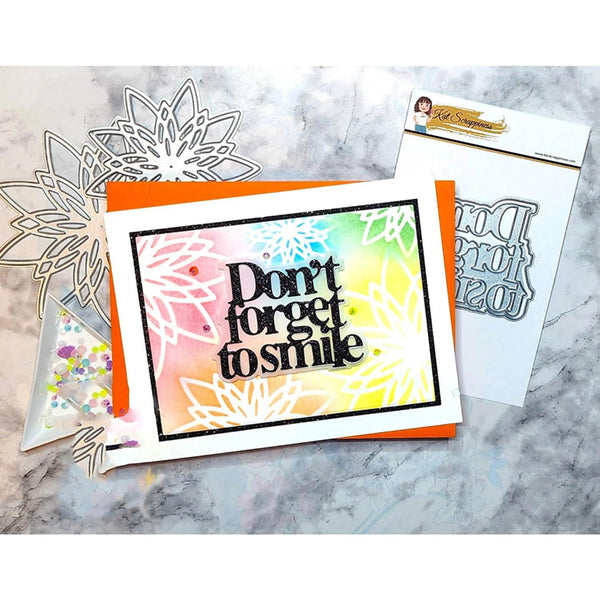 A handmade greeting card with the phrase 'Don't forget to smile' in black script over a pastel rainbow and floral pattern, displayed with metal die-cut embellishments and a stamp sheet on a gray marble surface.