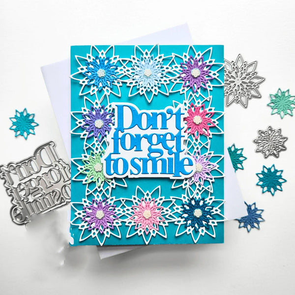 A handmade layered card with a turquoise background, white scalloped snowflake borders, and colorful flower accents around the words 'Don't forget to smile' in blue and white. The card is photographed on a white surface with metal die-cut embellishments and scattered snowflake pieces around it.