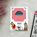 A square white card featuring circus-themed illustrations, including red-and-white vertical stripes, a blue sign reading "CIRCUS," and cartoon animals in clown outfits, placed on a textured light gray surface with part of a clear stamp sheet visible at the right edge.