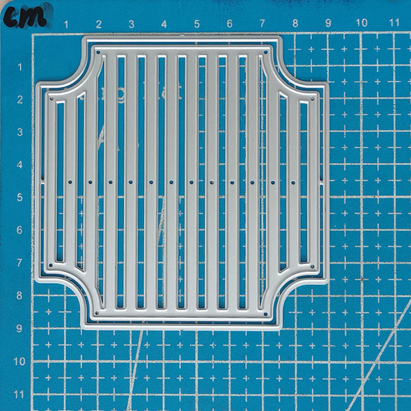 A silver metal die-cutting or stamping template with a decorative arched border and vertical slotted center, shown on a blue grid cutting mat.