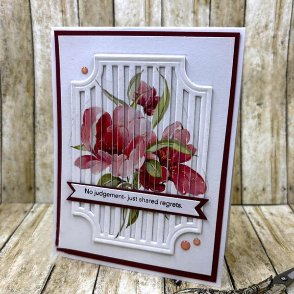 A handmade greeting card with a white and dark red layered design, featuring a floral motif in pink and red with green leaves. A banner across the lower middle reads 'No judgement - just shared regrets.