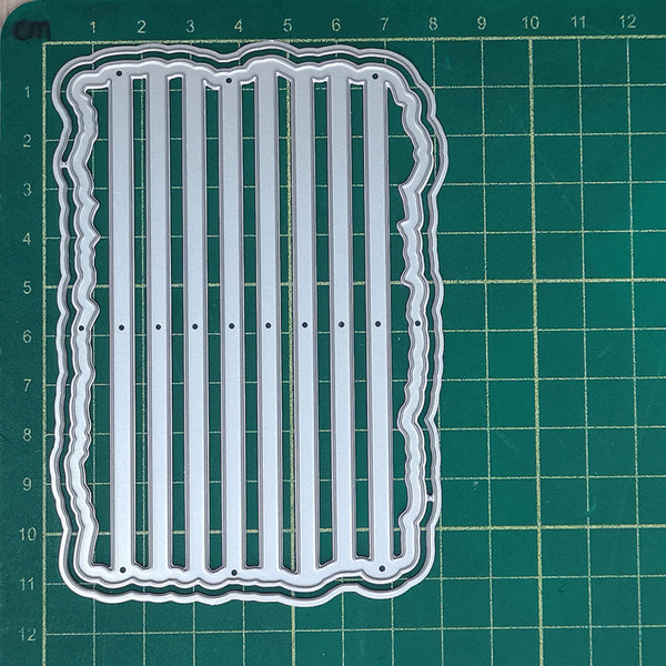 A clear acrylic or plastic embosser or stamping plate with vertical slits and a wavy border, placed on a green cutting mat with measurement markings.