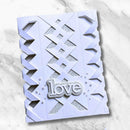 A handmade paper card with a pale lavender background, featuring a geometric lattice design with glittery silver cutouts and a glittery silver 'love' embellishment, displayed on a light gray marble surface.