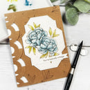A handmade greeting card with a brown kraft paper base, laser-cut scalloped edges, and a central white octagonal label featuring a blue peony illustration with yellow-green leaves. The card is placed on a white wooden surface with a black pen, glasses, and green leaves nearby.