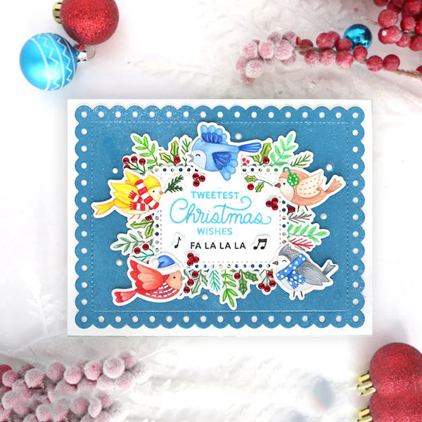 A square handmade Christmas greeting card with a scalloped blue border and colorful bird illustrations, photographed on a white surface with red and blue holiday ornaments around it.