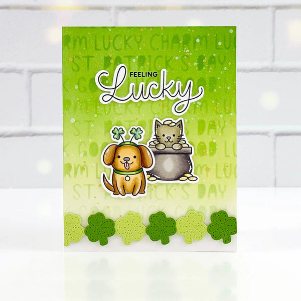 A St. Patrick's Day greeting card featuring a green gradient background with shamrock text, the words 'FEELING Lucky' in white script, a cartoon dog wearing a hat with three shamrocks, a cartoon cat sitting in a pot, and a row of die-cut shamrocks along the bottom edge.