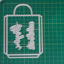 A silver metal die-cut craft template shaped like a shopping bag with the word 'Bigo' repeated vertically in script, placed on a green cutting mat with grid lines.