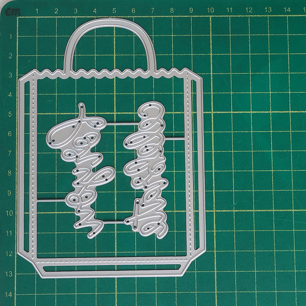 A silver metal die-cut craft template shaped like a shopping bag with the word 'Bigo' repeated vertically in script, placed on a green cutting mat with grid lines.