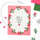 A handmade greeting card with a pink border and a white center featuring the text 'HAVE A JOYFUL DAY!' surrounded by a 3D floral wreath made of pink paper flowers and green leaves. The card is styled on a white background with craft cutouts and colored paper pieces.