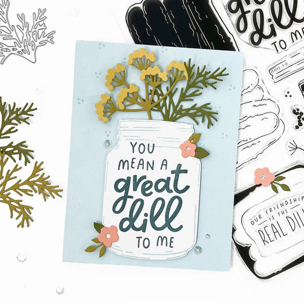 A handmade greeting card featuring a mason jar design with the phrase 'YOU MEAN A great dill TO ME' in dark teal script, decorated with yellow flowers and green foliage. The card is styled on a white background alongside other crafting elements, including clear stamps and gray die-cut embellishments.