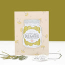 A handmade greeting card featuring an illustration of a glass jar filled with pickled cucumbers and the text 'HAVE A DILLIGHTH DAY'. The card is displayed on a white surface with a yellow-green background, accompanied by a small tag and sprigs of greenery.